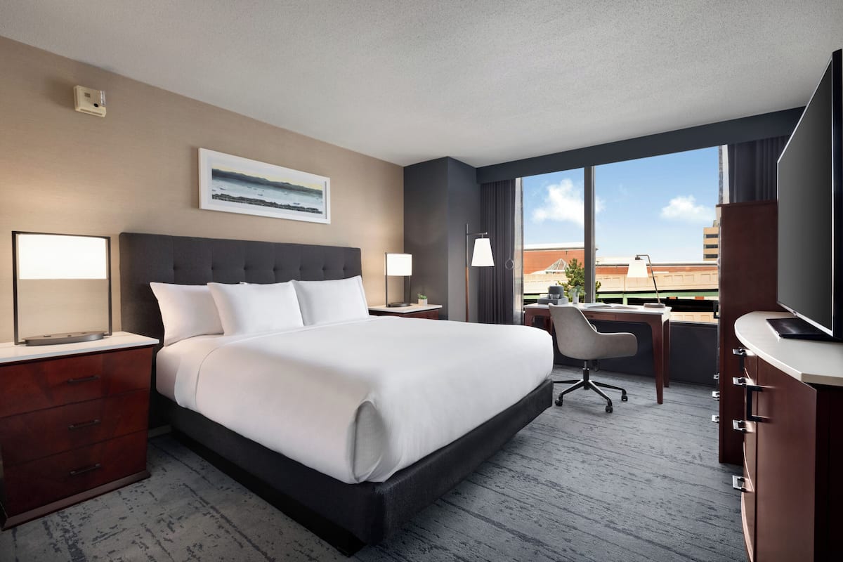 A hotel room with a large bed, a desk with a chair, a TV, and a window with a city view.
