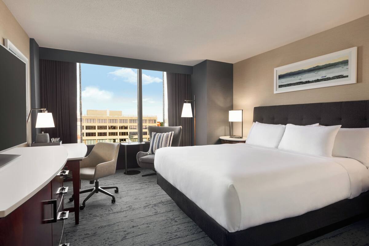 A hotel room with a large bed, a desk, a chair, and a view of the cityscape through the window.