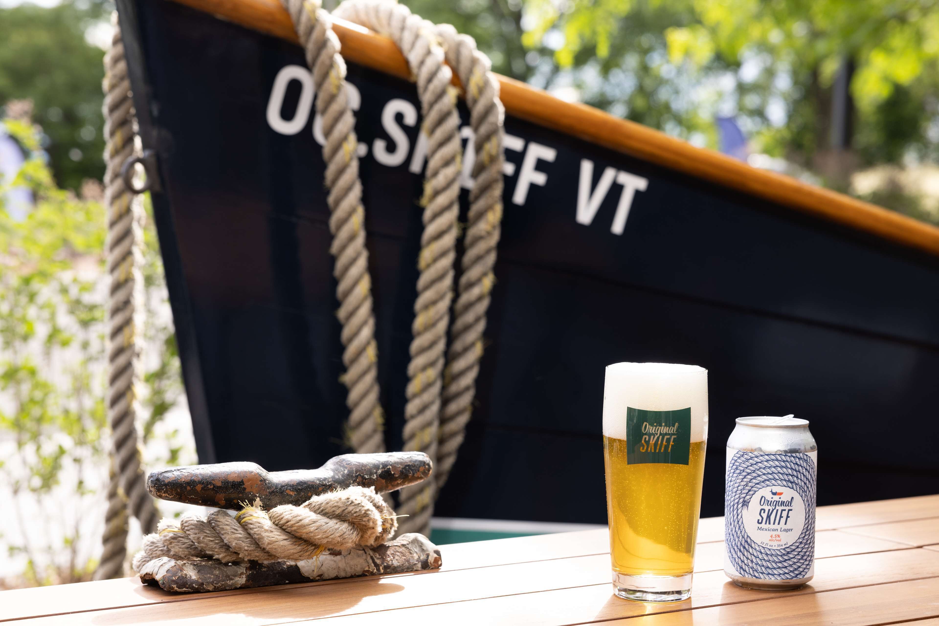 A wooden table with a glass of beer and a can of beer, a rope, and a boat in the background.