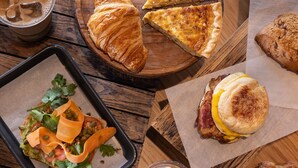 Una mesa de madera con distintos alimentos, como un sándwich, un croissant y una bebida.