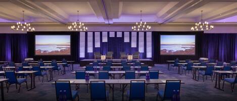 A conference room with multiple tables and chairs, large screens, and chandeliers.