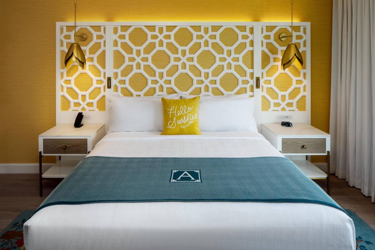 A hotel room with a modern design, featuring a geometric headboard, a bed with a teal bedspread, and a yellow pillow with text.