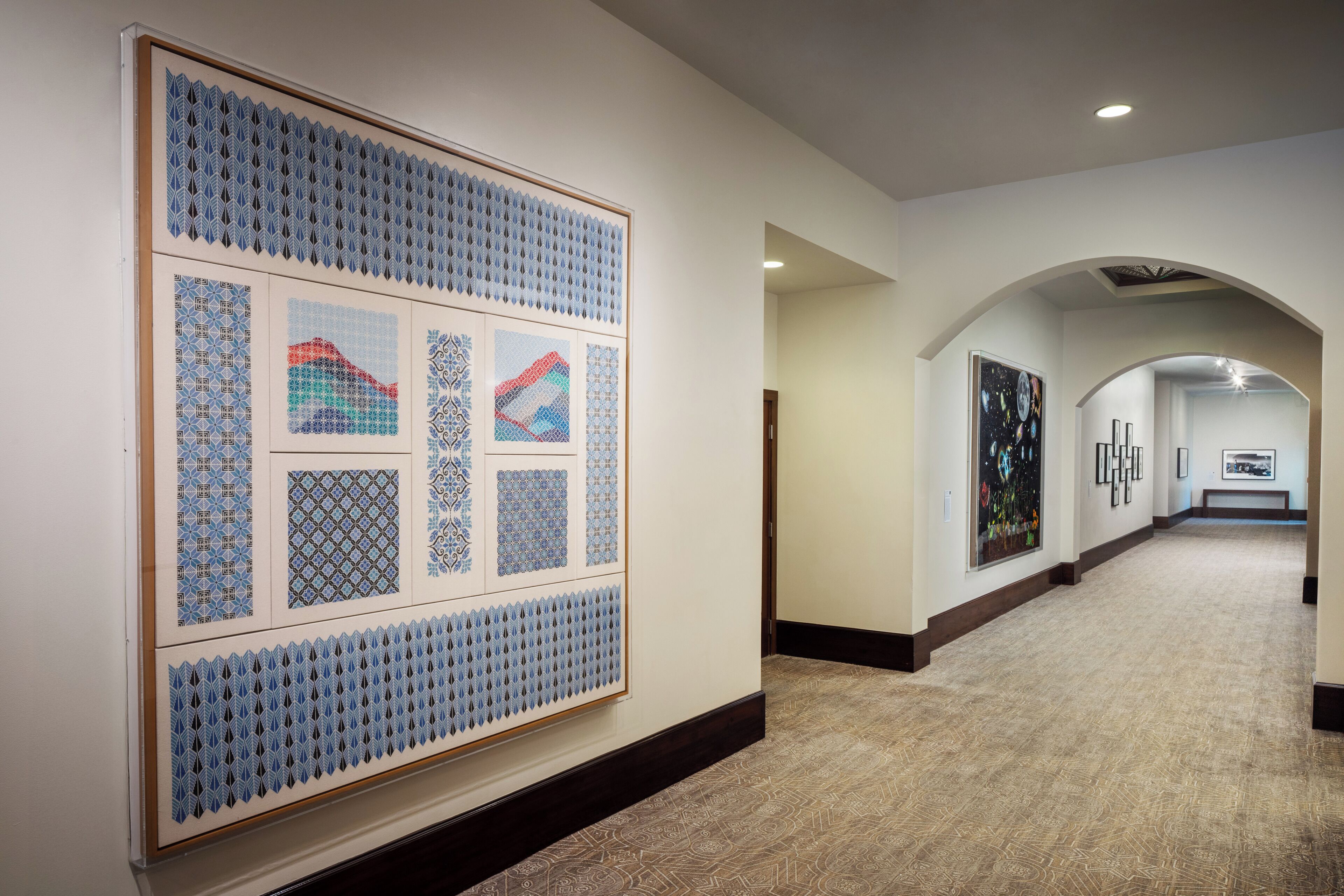 A hallway with a large framed artwork on the left, a patterned carpet, and a view of the room in the distance.