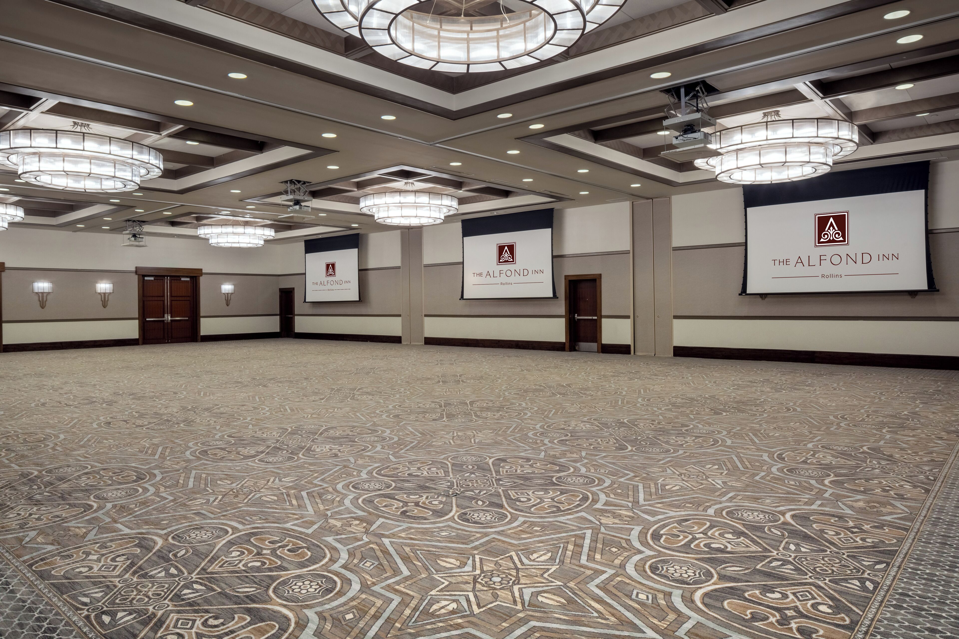 A spacious hotel lobby with patterned carpet, multiple chandeliers, and banners for The Alfond Inn.