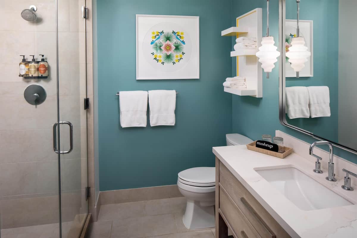 A modern bathroom with a shower, toilet, sink, and a mirror. There are towels on the wall and a framed artwork above the toilet.