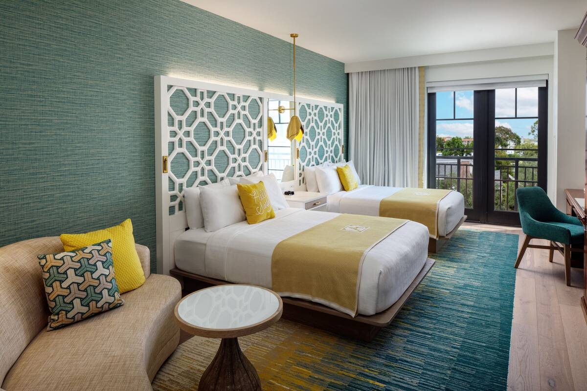 A hotel room with two beds, a sofa, a small table, and a balcony with a view.