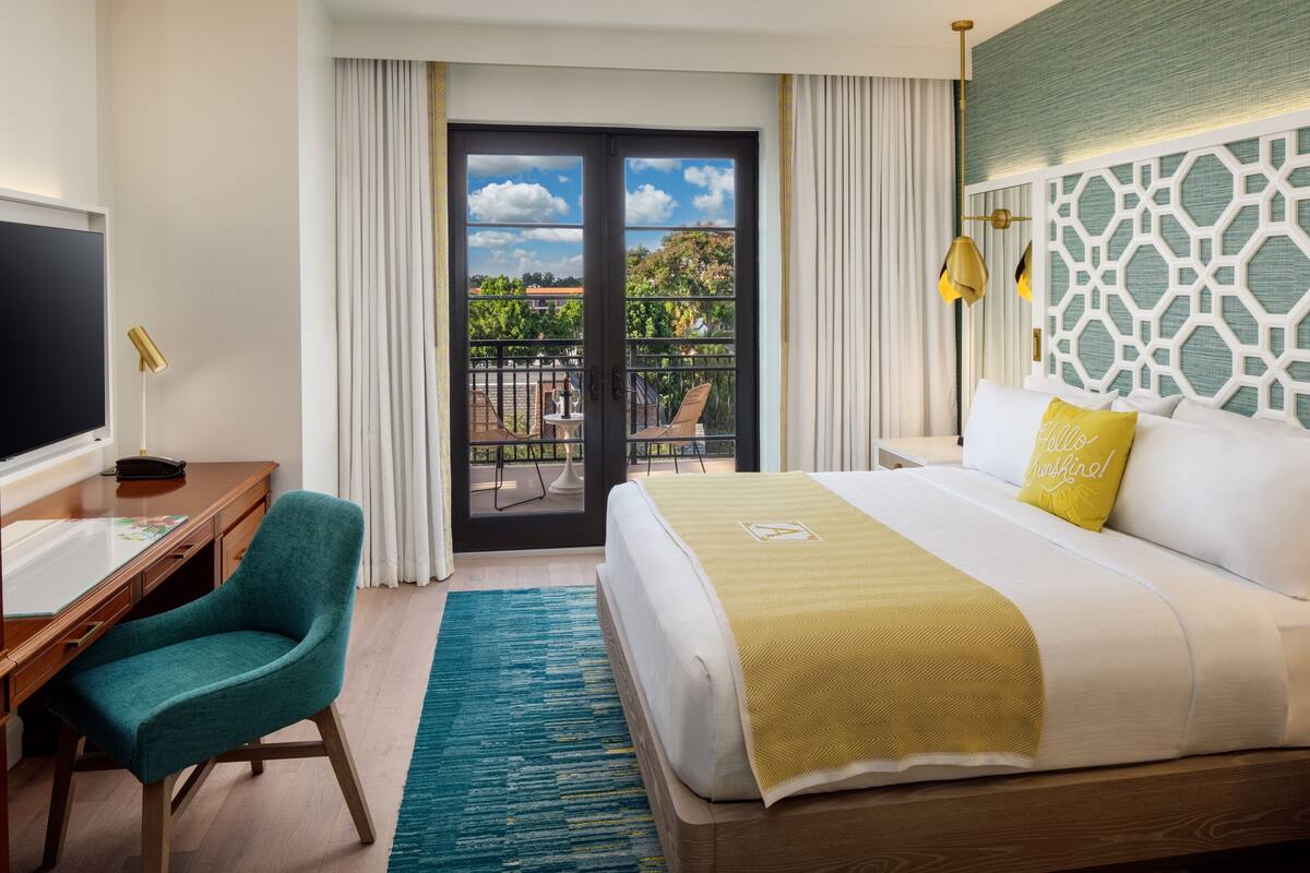 A hotel room with a bed, a desk, a chair, a balcony with a view, and a TV.