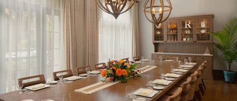 A long dining table set for a meal with a centerpiece, surrounded by chairs, in a room with wooden flooring and a cabinet with glass doors.