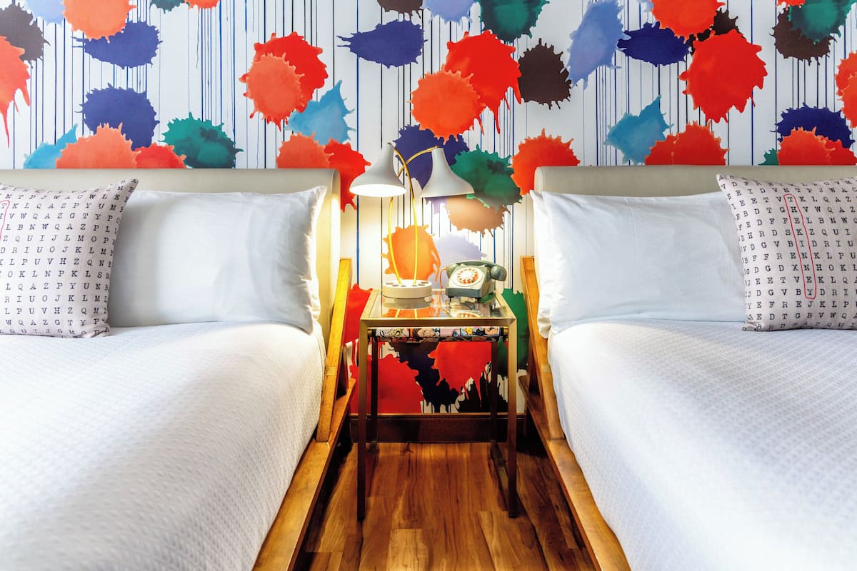 A room with two beds, a nightstand with a lamp, and a wall with colorful splatter patterns.