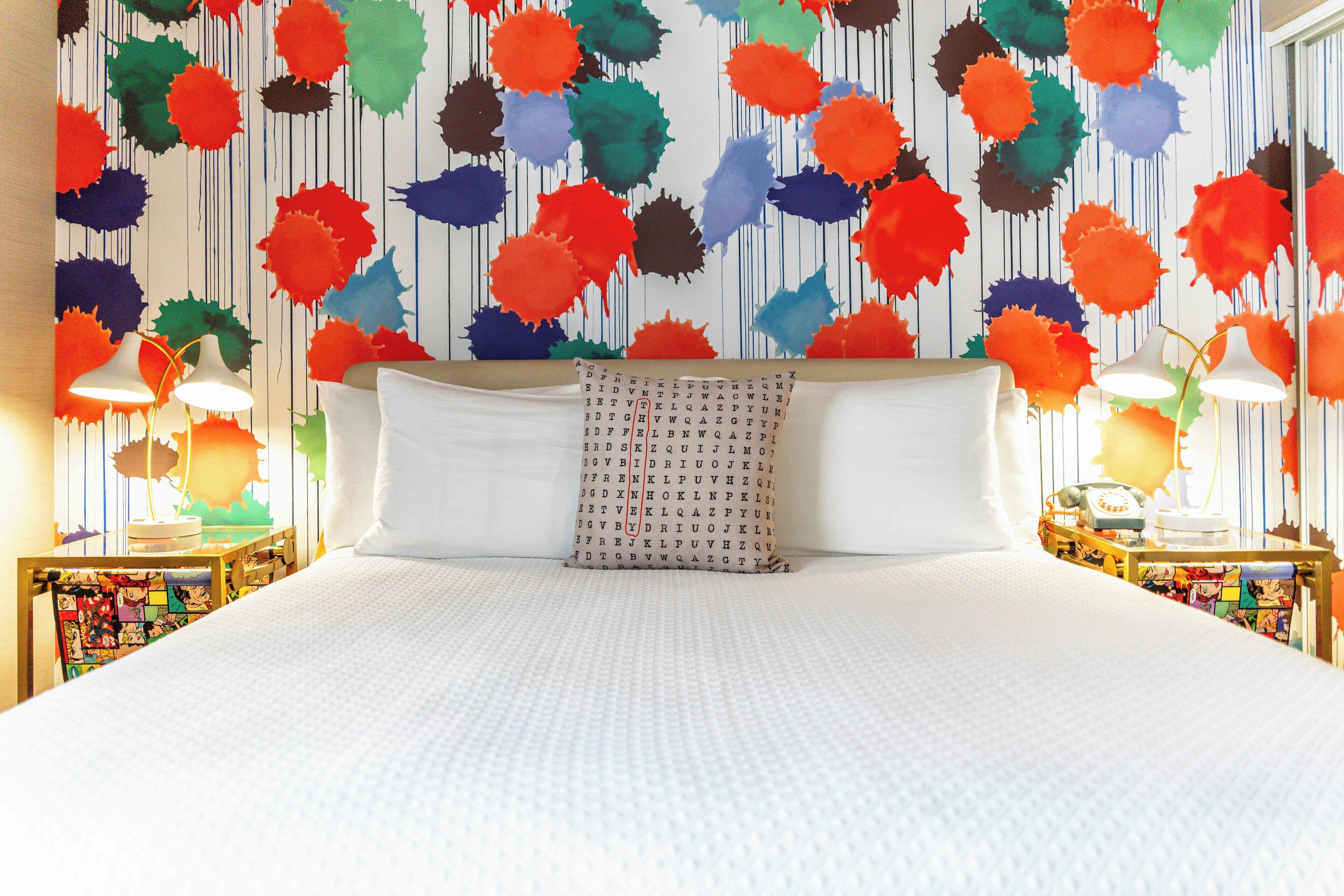 A bed with a white duvet, a pillow with a pattern, and a wall with colorful splatter designs. There are two bedside tables with lamps.