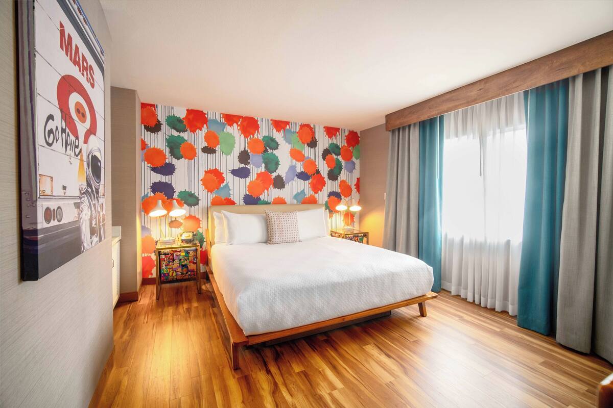 A hotel room with a wooden floor, a bed with white bedding, a colorful wall mural, and a window with curtains.