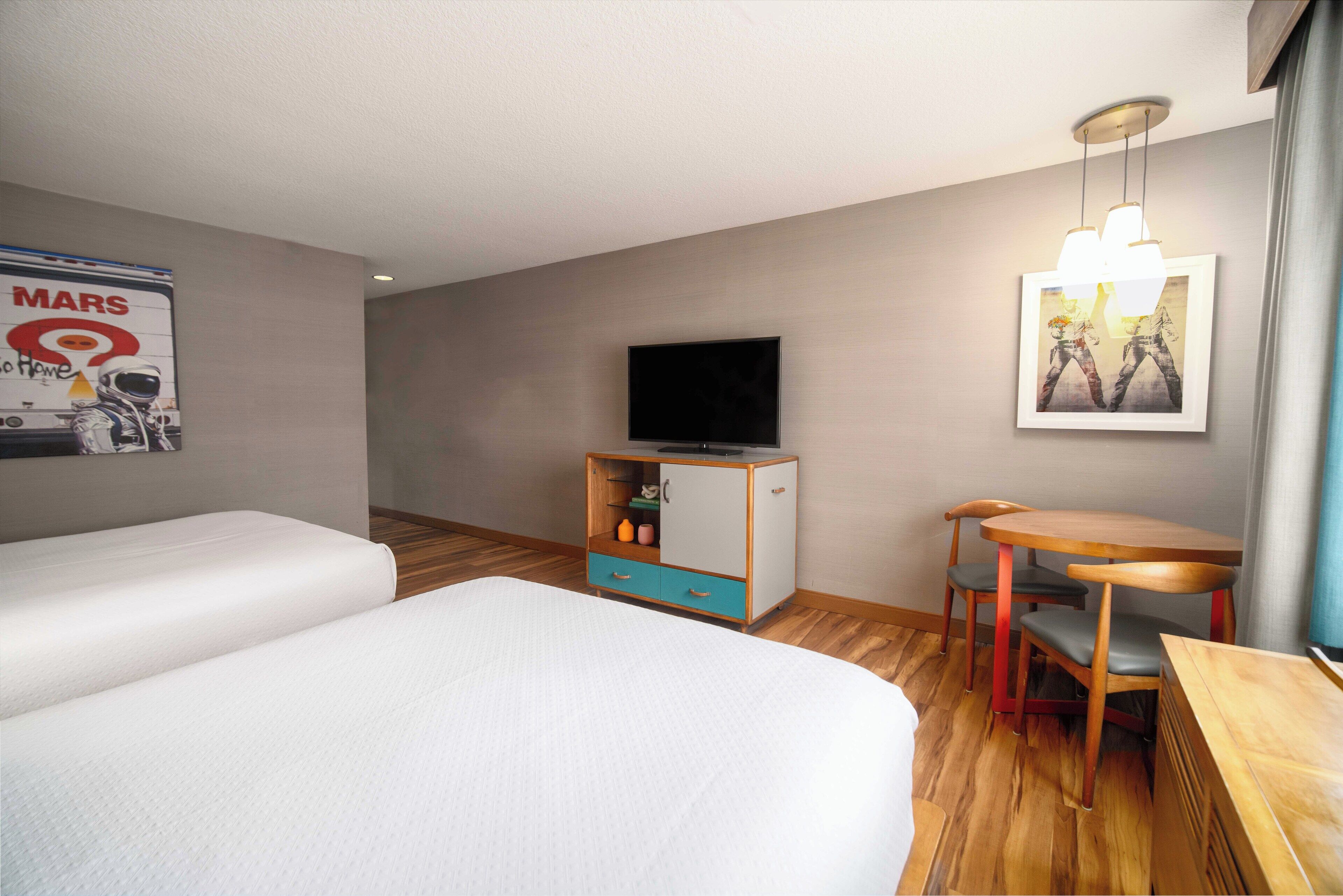 Ein Hotelzimmer mit zwei Betten, einem Fernseher, einem kleinen Tisch mit Stühlen und einer Kommode mit Schubladen.