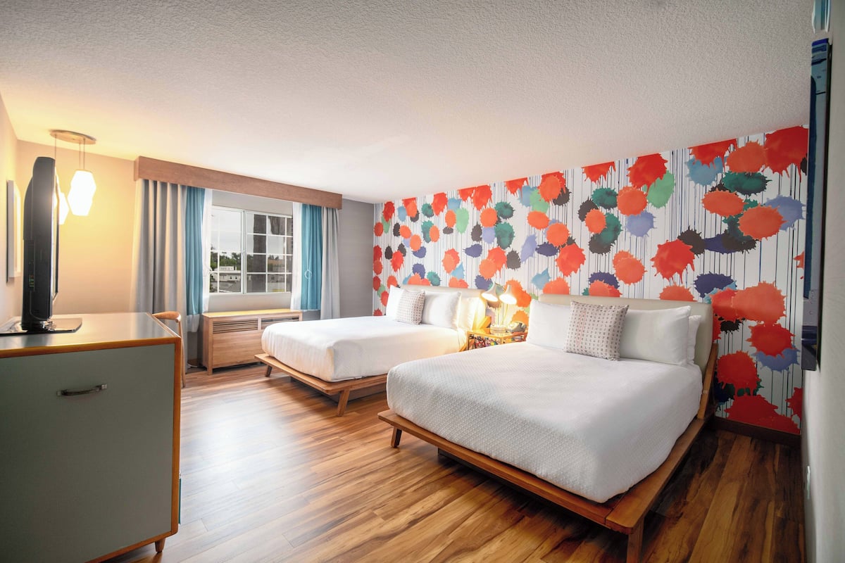 A hotel room with two beds, a desk, a TV, and a window with curtains.
