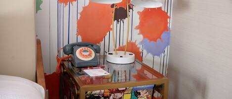 A hotel room with a bed, a wooden nightstand with a telephone, a lamp, and a colorful comic book-themed table.