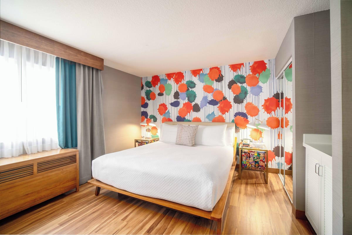 A modern bedroom with a wooden bed, a window with curtains, a colorful wall mural, and a nightstand with a lamp.