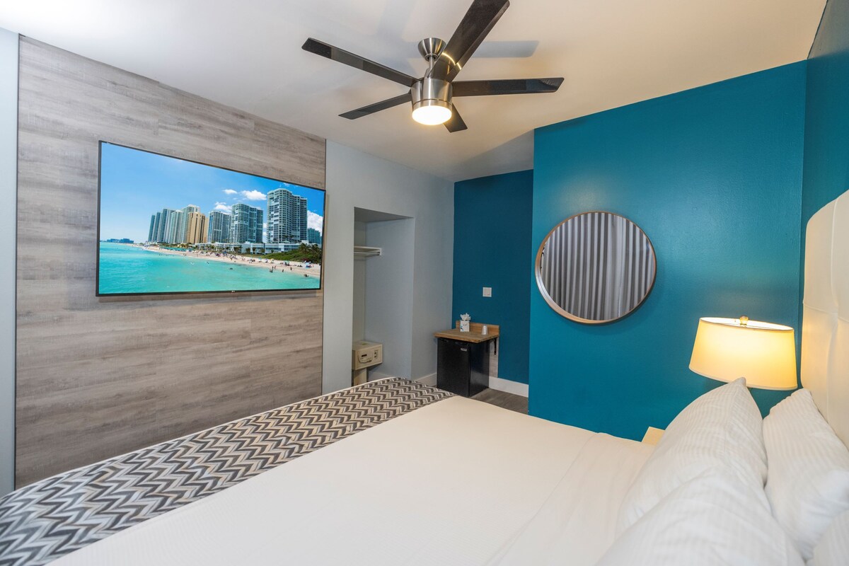 A hotel room with a bed, a ceiling fan, a TV displaying a beach scene, a small bedside table, and a wall-mounted lamp.