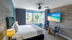 Ein Hotelzimmer mit Bett, Fernseher, Deckenventilator, einem Fenster mit Vorhängen und Blick auf einen Strand.