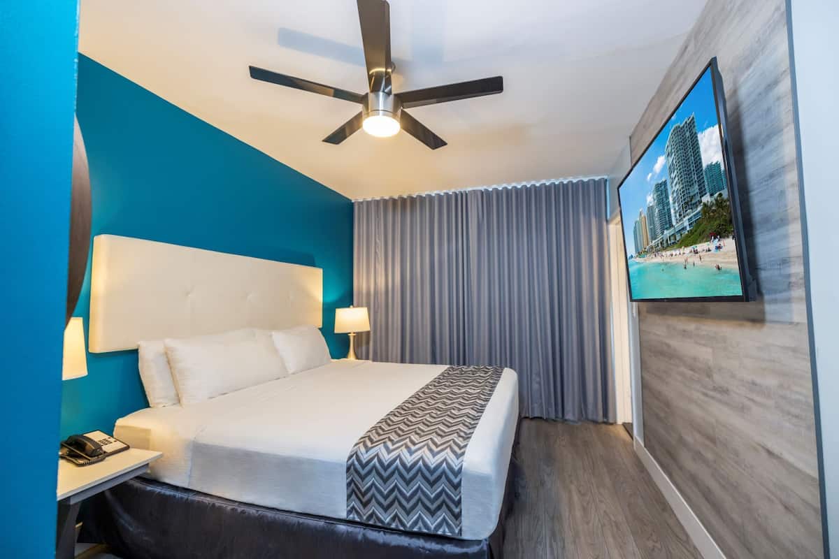 A hotel room with a bed, a desk with a telephone, a ceiling fan, and a large window with curtains.