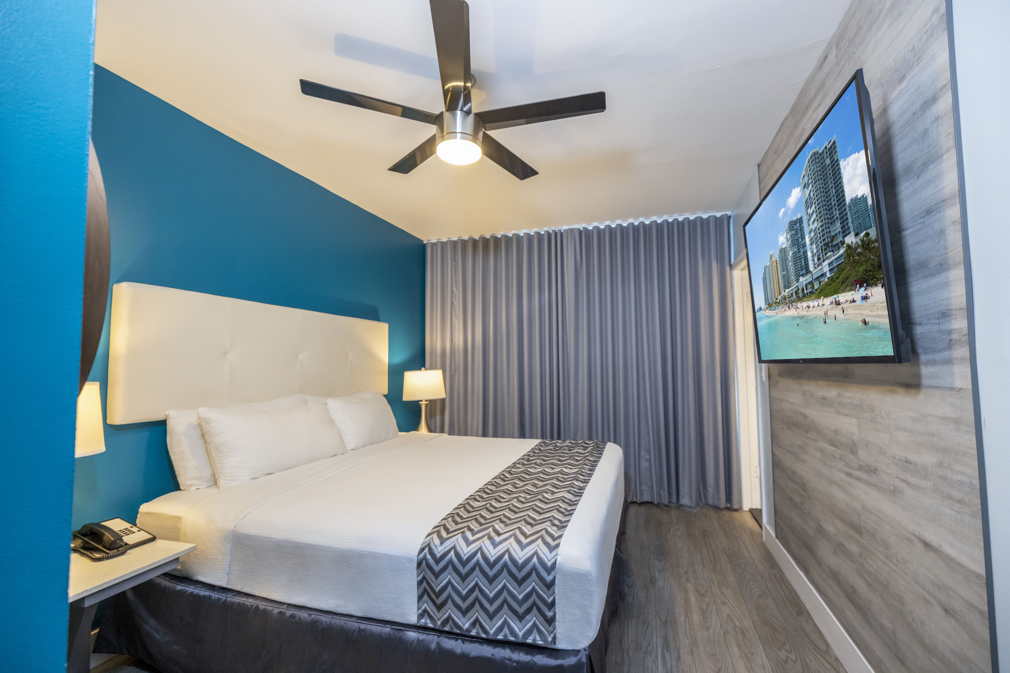 A hotel room with a bed, a desk with a phone, a ceiling fan, and a TV displaying a beach scene.