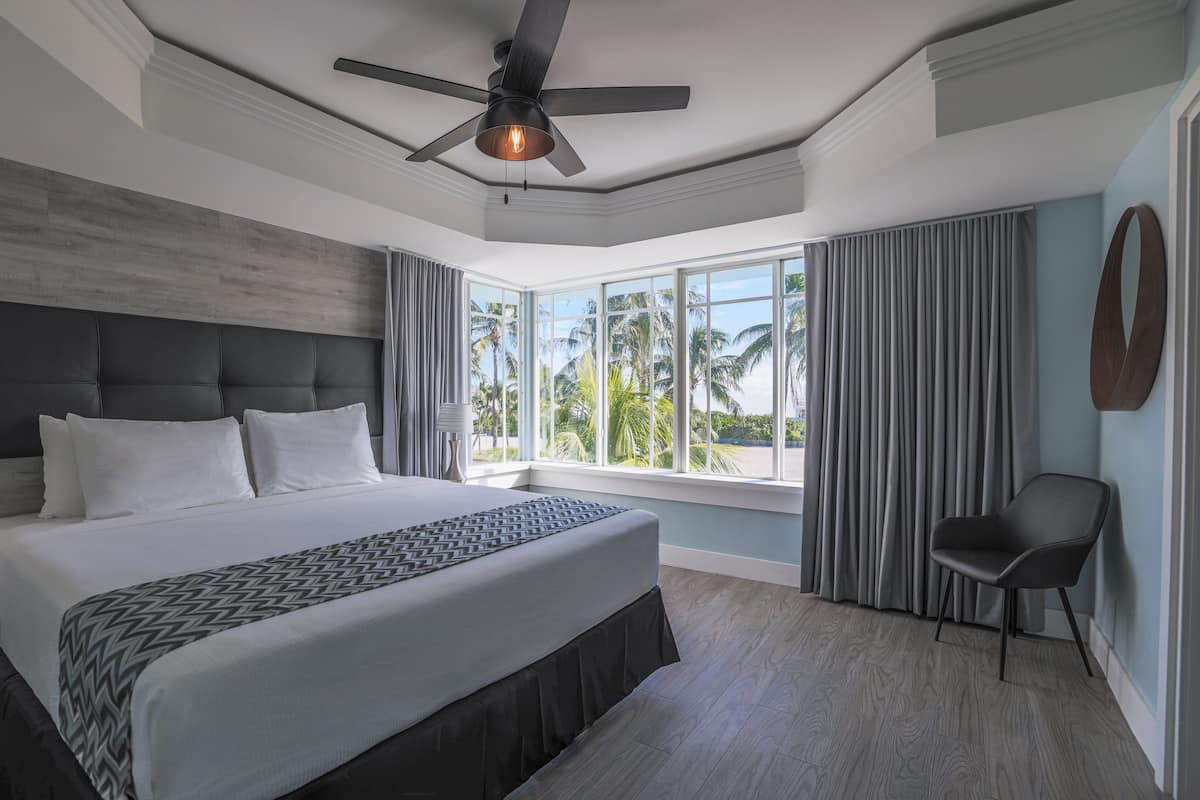A modern hotel room with a large bed, a ceiling fan, a view of palm trees, and a chair.