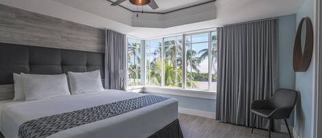 A modern hotel room with a large bed, a ceiling fan, a view of palm trees, and a chair.