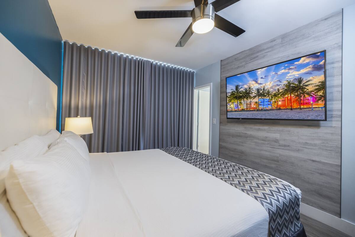 A hotel room with a bed, a ceiling fan, a TV on the wall, and a window with curtains.