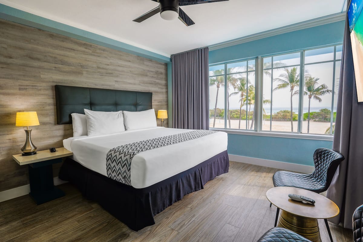 A hotel room with a large bed, a ceiling fan, a view of palm trees, and a small table with a remote control.