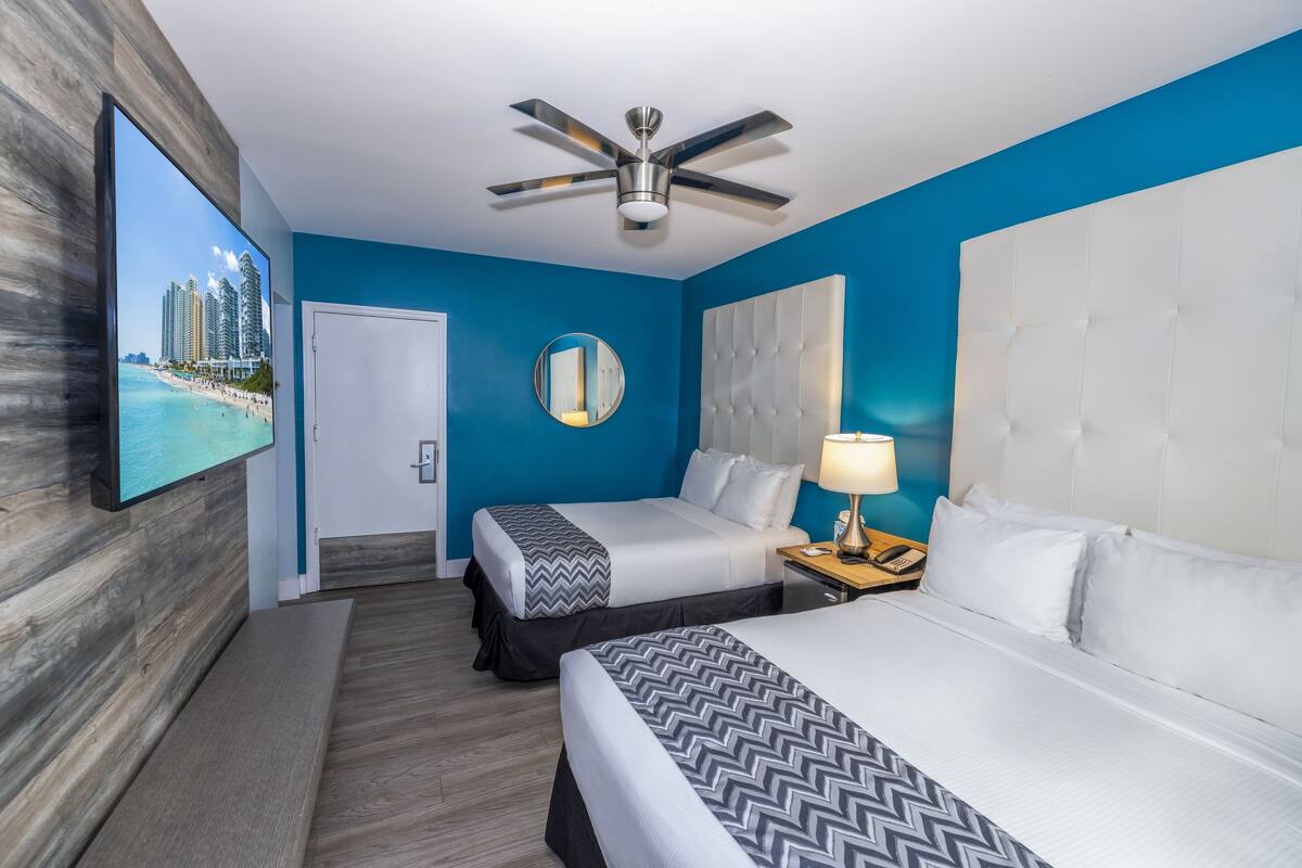 A hotel room with two beds, a ceiling fan, a round mirror, and a TV displaying a beach scene.