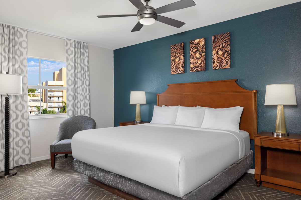 A hotel room with a large bed, two bedside tables with lamps, a chair, a ceiling fan, and a window with curtains.