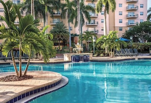 A swimming pool with a fountain, surrounded by palm trees and lounge chairs, with a multi-story building in the background. - Club Wyndham Palm-Aire (Pompano Beach)
