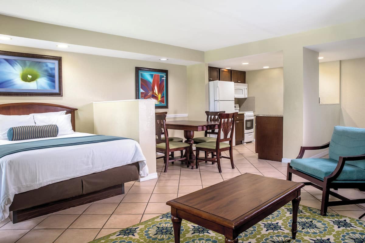 A hotel room with a bed, a dining table with chairs, a kitchen area, and a seating area with a chair.