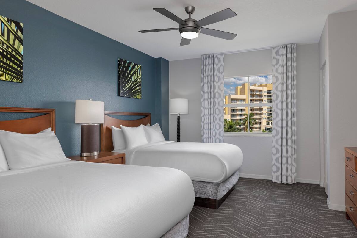A hotel room with two beds, a ceiling fan, a window with patterned curtains, and artwork on the walls.
