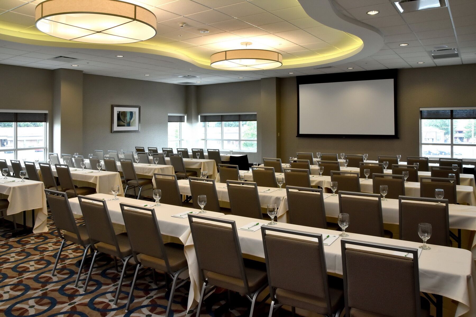 Un salón de conferencias con mesas dispuestas para una reunión, una pantalla de proyector y un ventanal grande.