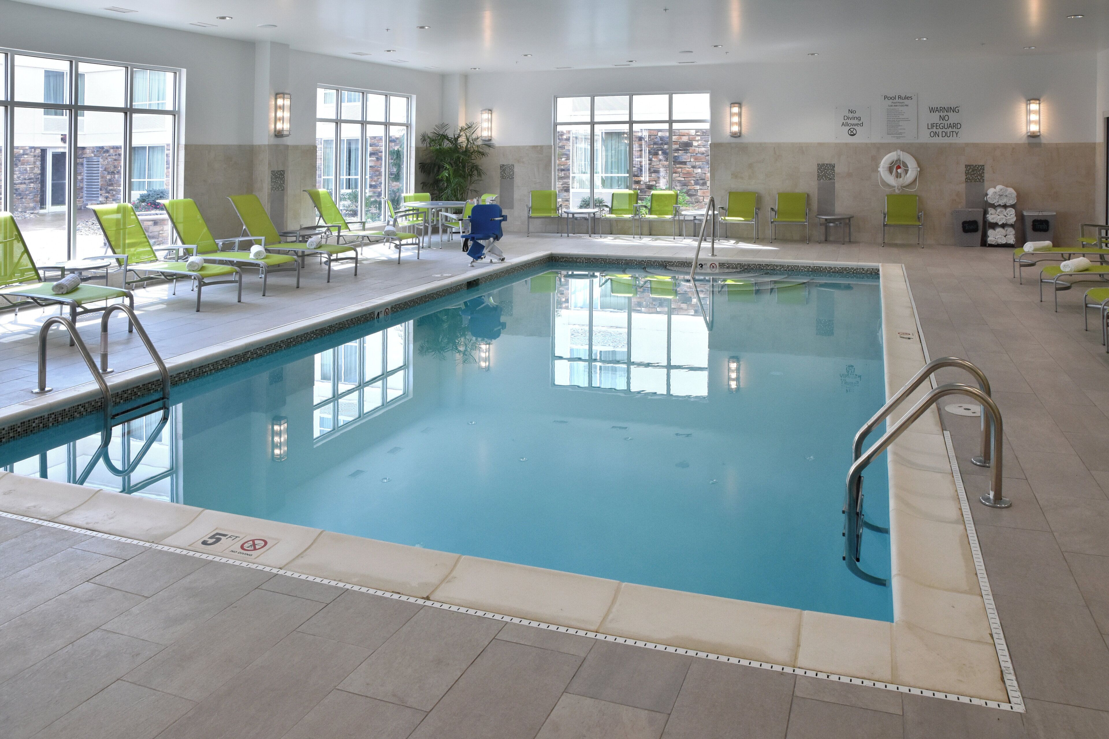 An indoor swimming pool with clear water, surrounded by green lounge chairs and a tiled floor.
