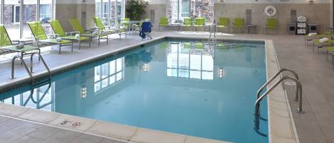 Una piscina interior con agua cristalina, rodeada de sillas reclinables verdes y suelo de baldosas.