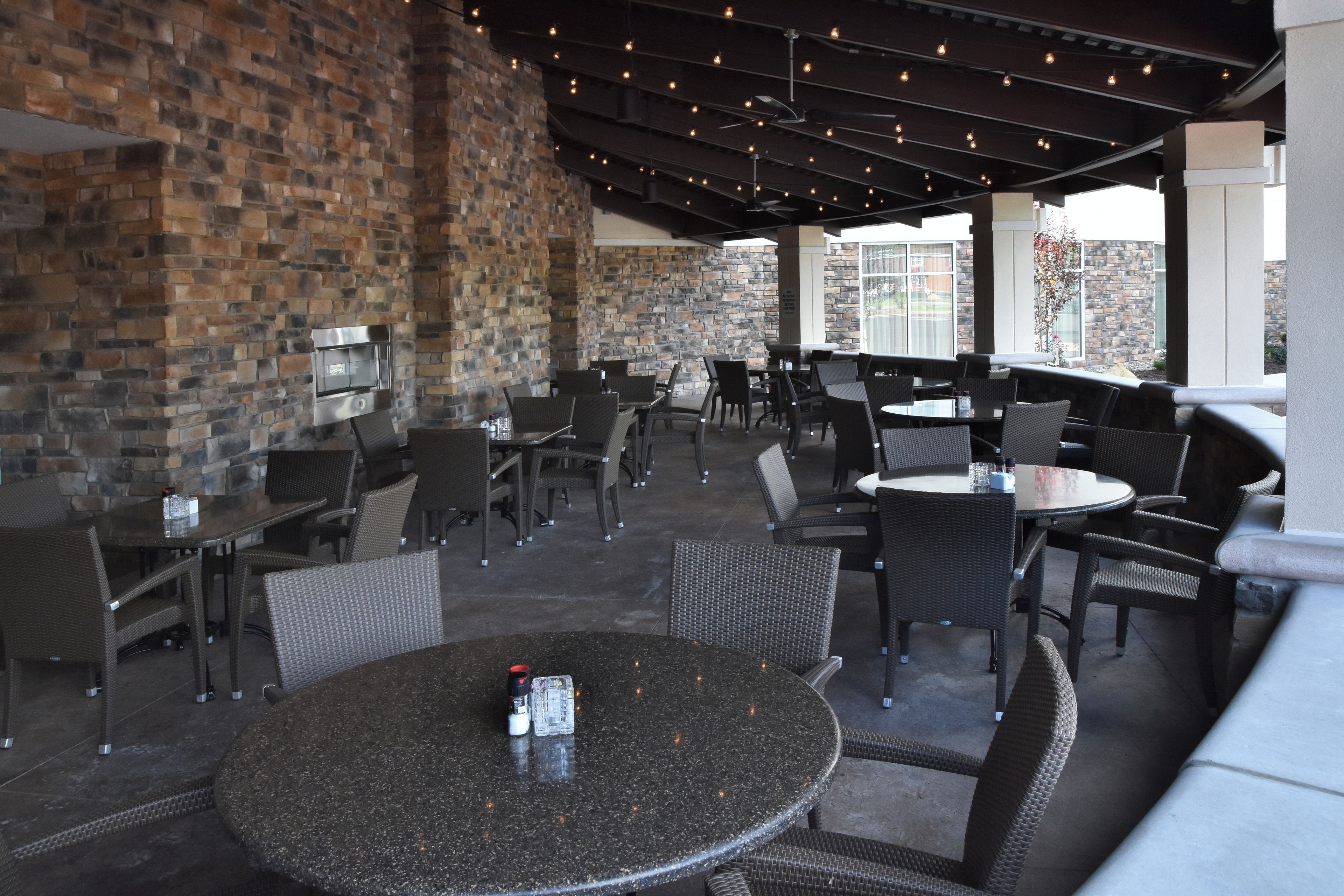 An outdoor patio area with brick walls, multiple seating arrangements including round tables and chairs, and a ceiling with exposed beams and lighting.
