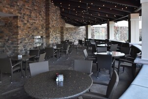 An outdoor patio area with brick walls, multiple seating arrangements including round tables and chairs, and a ceiling with exposed beams and lighting.