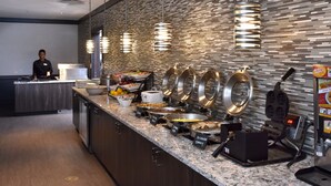 A breakfast buffet with various food stations, a person at the counter, and a waffle maker. - Holiday Inn Canton (Belden Village) by IHG (Canton)