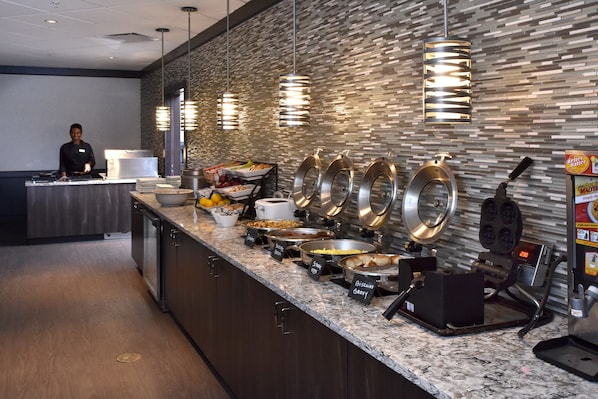 A breakfast buffet with various food stations, a person at the counter, and a waffle maker.