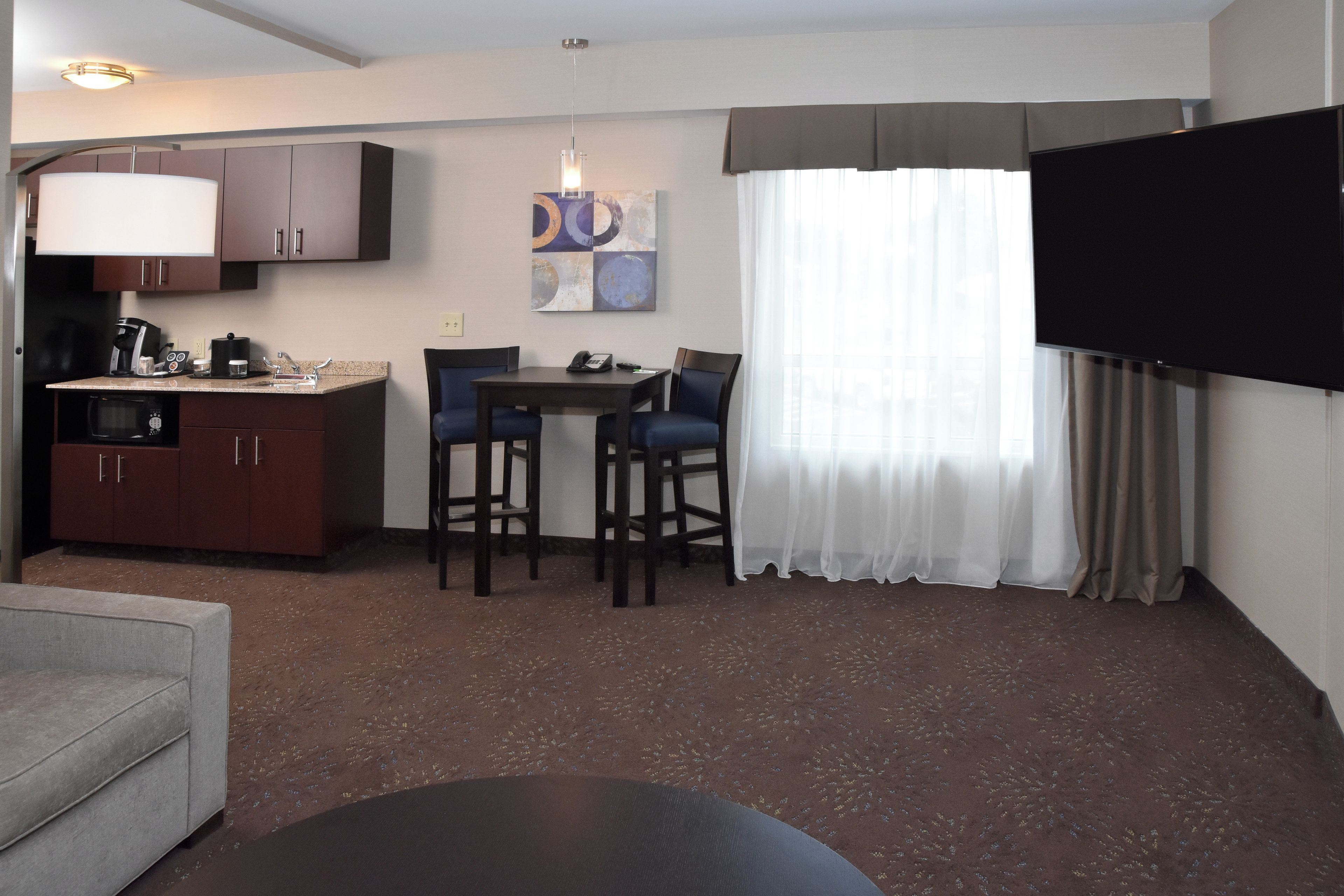 A compact hotel room with a kitchenette, a dining area with two chairs, a flat-screen TV, and a large window with sheer curtains.