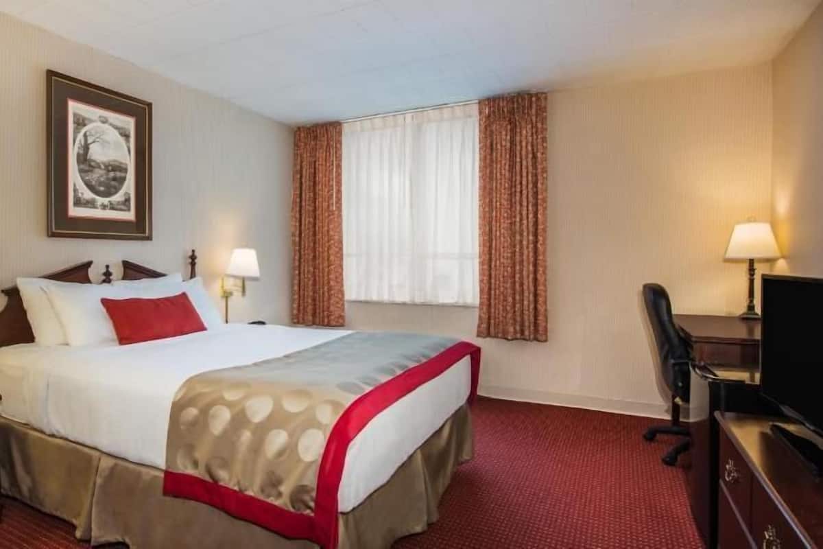 Ein Hotelzimmer mit einem Bett, zwei Nachttischen mit Lampen, einem Schreibtisch mit Stuhl, einem Fernseher und einem Fenster mit Vorhängen.