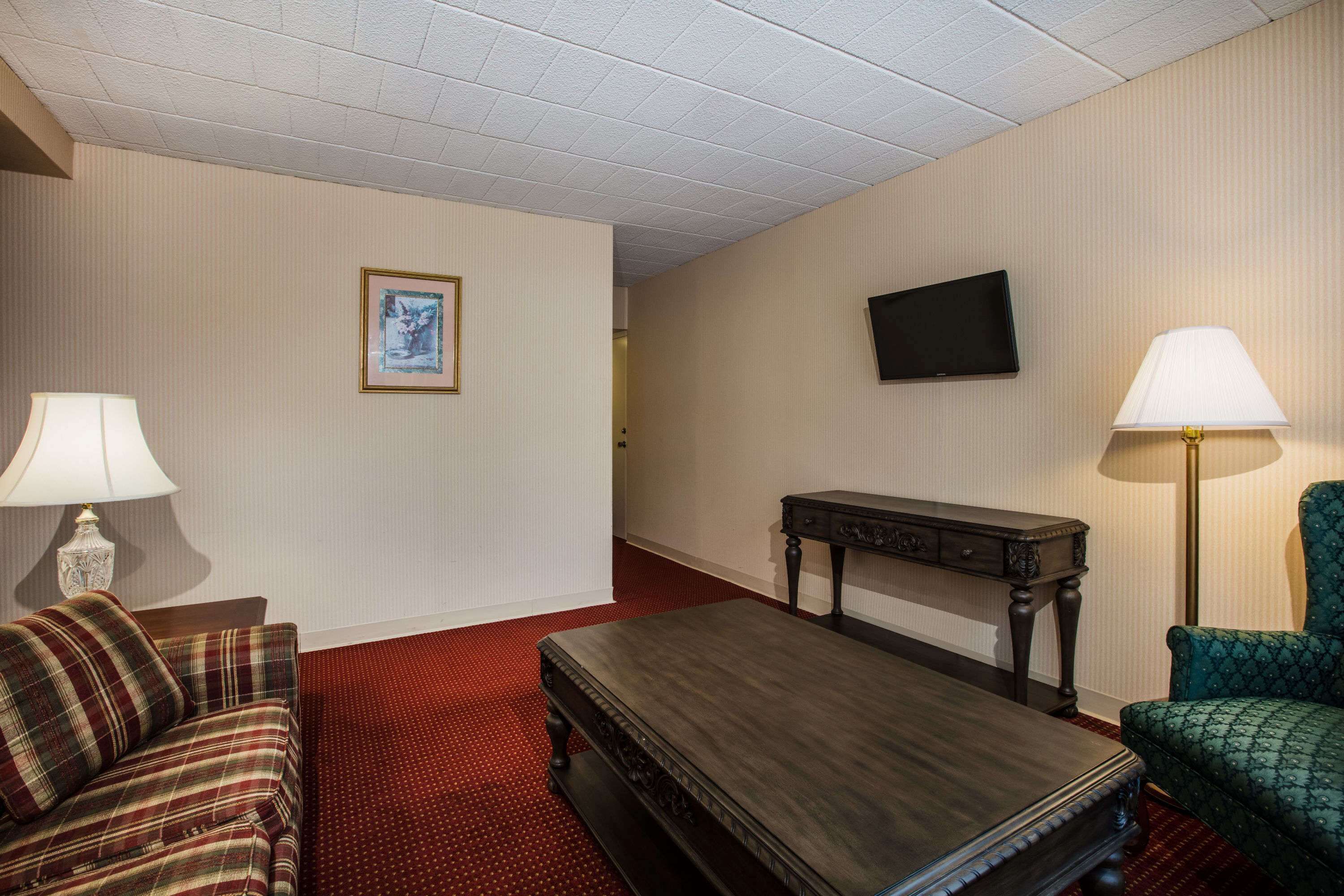 A hotel room with a plaid sofa, a dark wooden coffee table, a green armchair, a small side table, a flat-screen TV mounted on the wall, and a red carpeted floor.