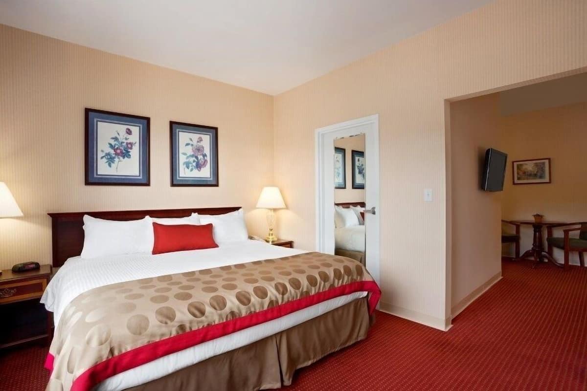 A hotel room with a large bed, two bedside lamps, a red throw pillow, a mirror, and a small table with chairs.