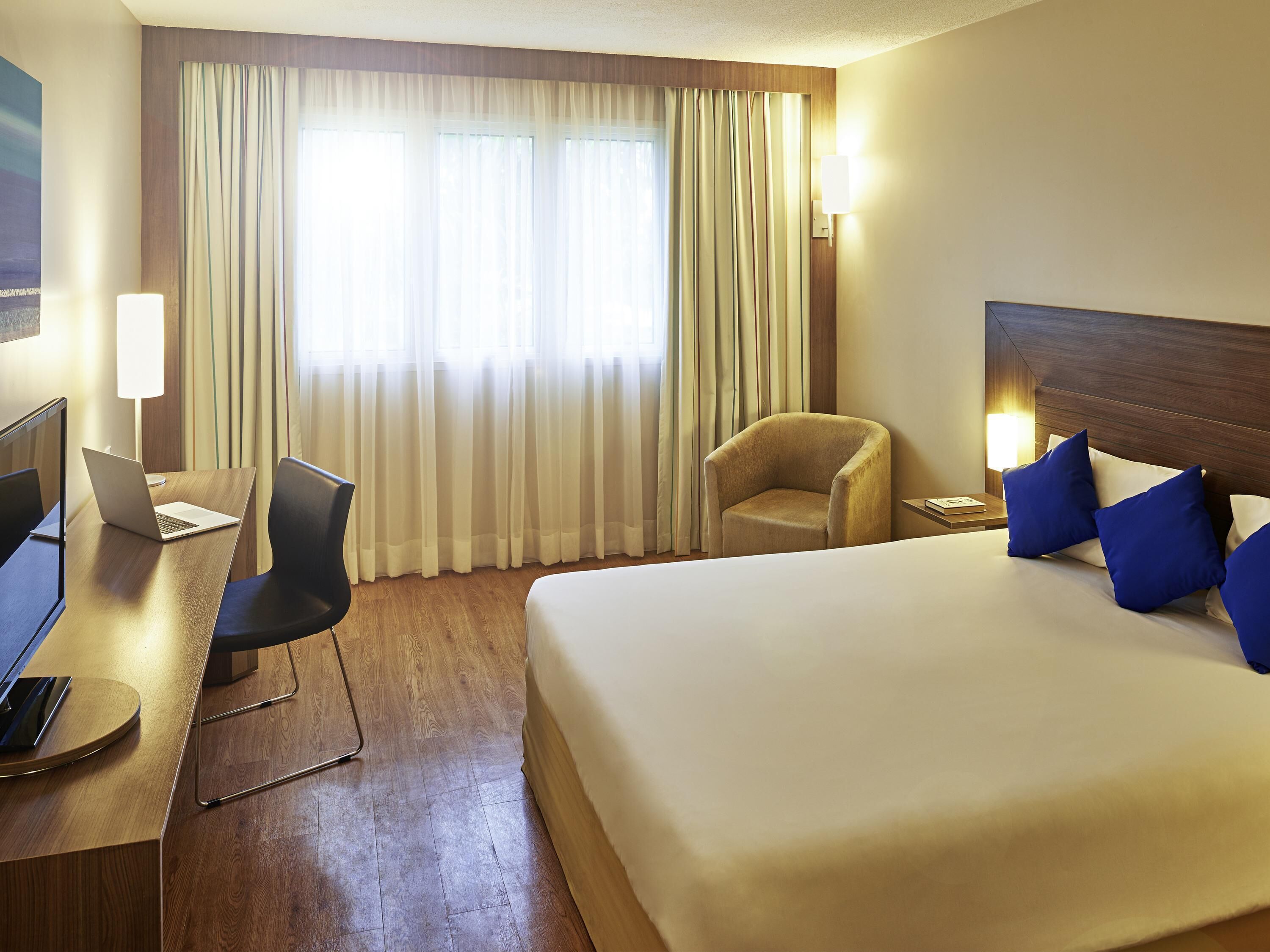 A hotel room with a bed, a desk with a laptop, a chair, and a window with curtains.