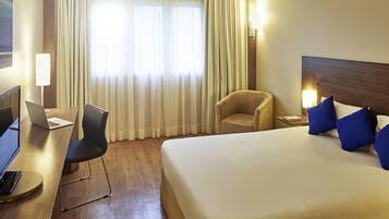 A hotel room with a bed, a desk with a laptop, a chair, and a window with curtains.