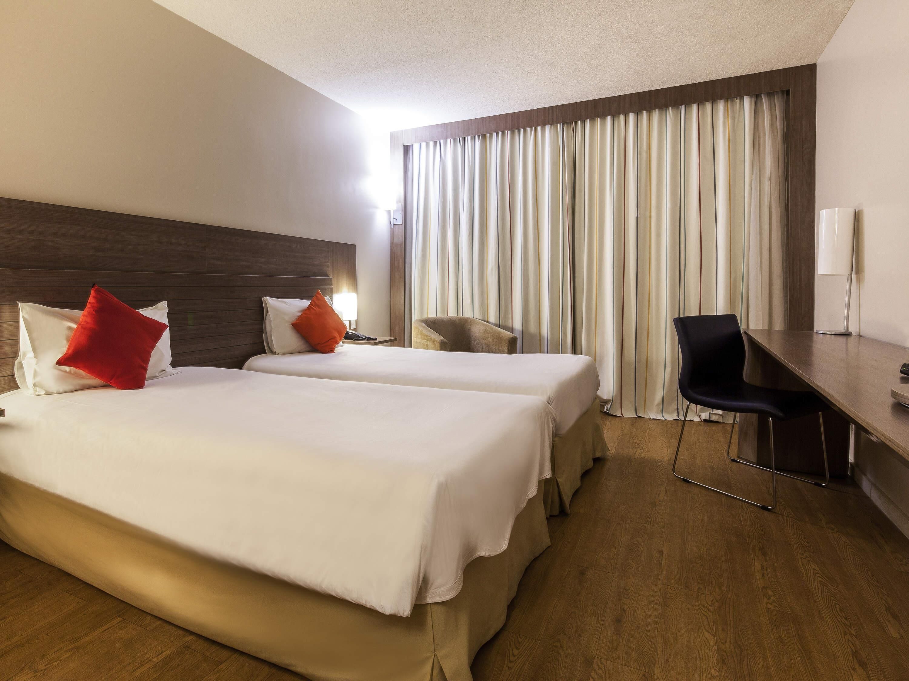 A hotel room with two beds, a desk, a chair, and a large window with curtains.