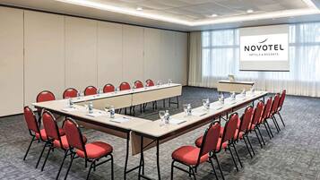 A conference room with a long table, red chairs, and a projector screen displaying "NOVOTEL HOTELS & RESORTS".