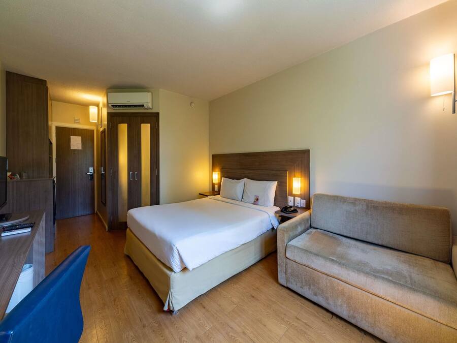 A hotel room with a bed, a sofa, a desk, and an air conditioning unit.