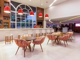 Un área de bar moderna con un ventanal grande, lámparas colgantes rojas y diversas opciones de asiento, incluyendo mesas redondas y sillas con diseños originales.