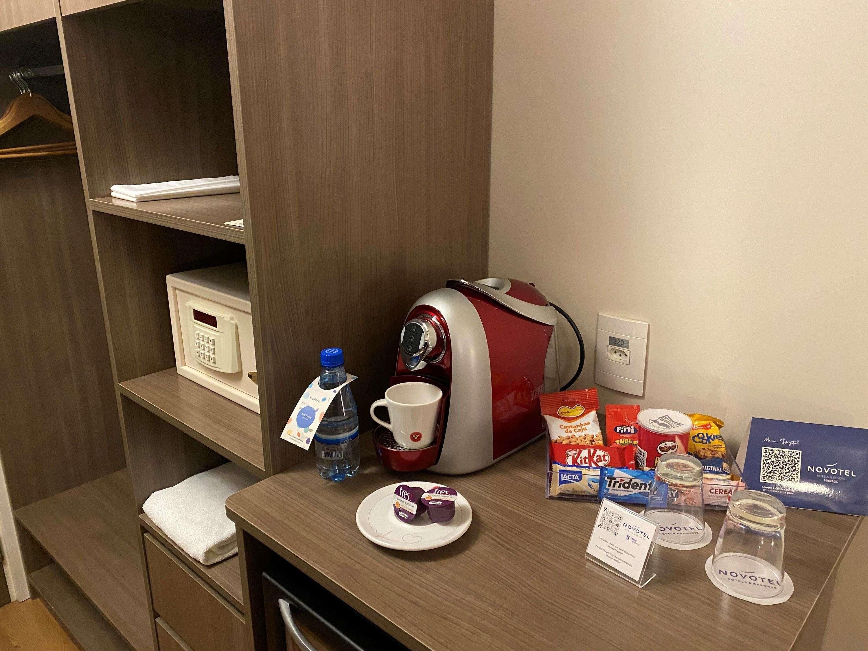 A hotel room with a kitchenette, a water bottle, a coffee maker, a plate with a dessert, and various food items on the counter.